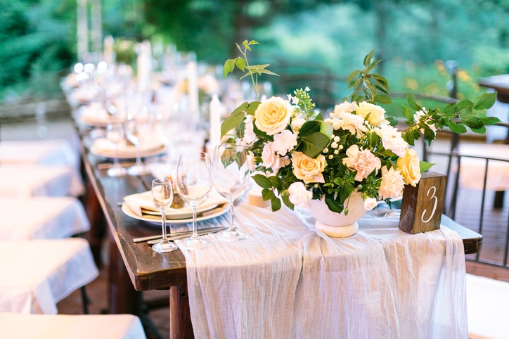 Festive outdoor wedding celebration table covered with natural tablecloth and beautifully neatly served with sets of dishes, glasses, appliances and flower arrangements on a green background intimate wedding
